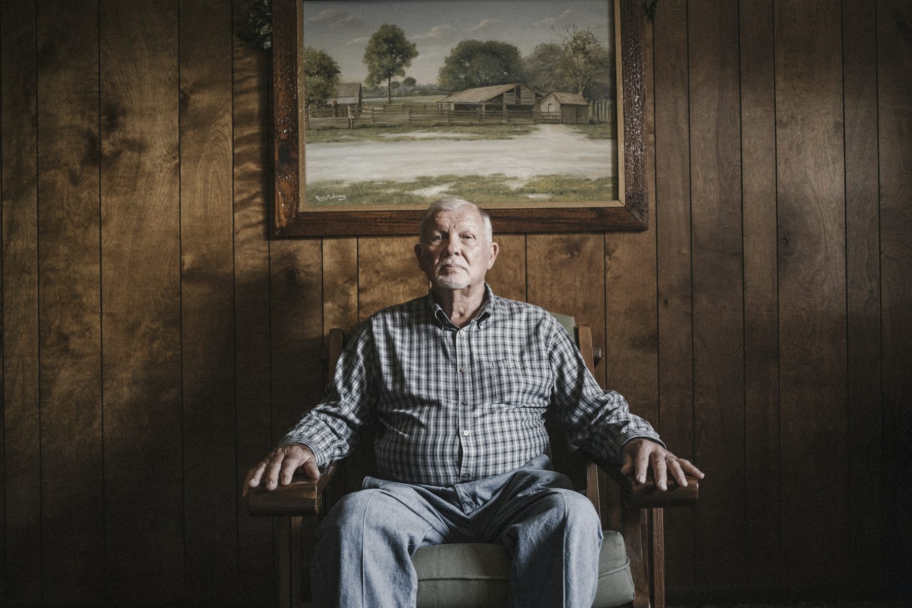 An older man sits in a wooden armchair beneath a landscape painting in a wood-paneled room